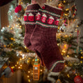 A hand is holding a pair of hand knitted socks in front of a Christmas tree. The colourwork pattern has candy cane stripes and cups of hot chocolate on them.