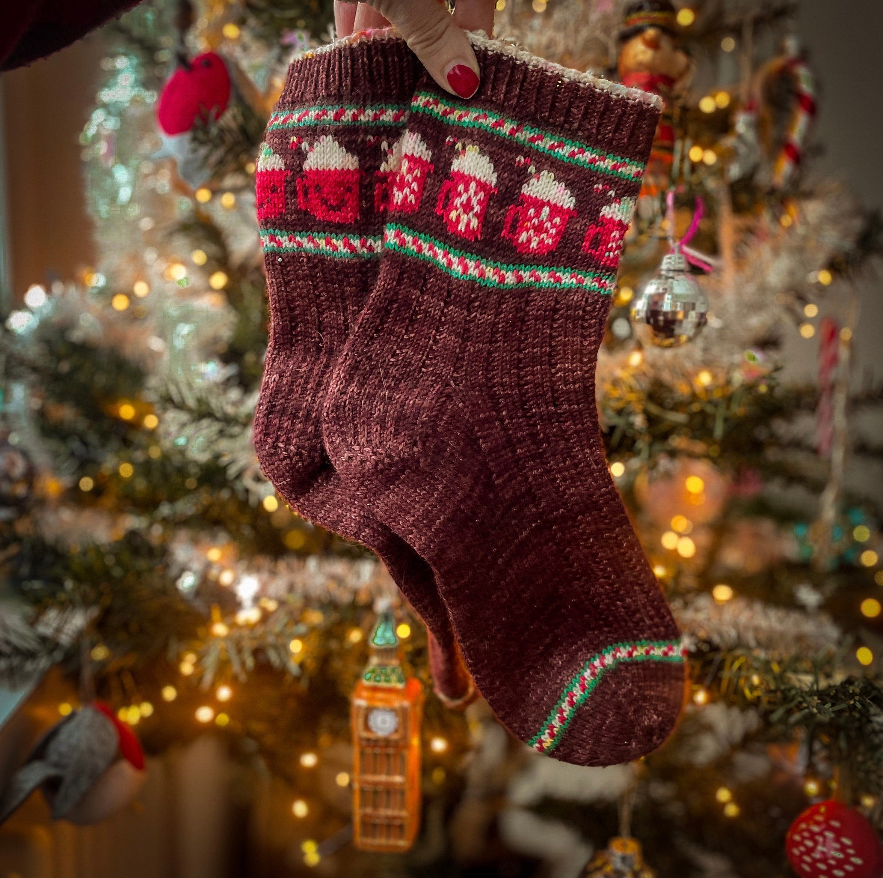 A hand is holding a pair of hand knitted socks in front of a Christmas tree. The colourwork pattern has candy cane stripes and cups of hot chocolate on them.